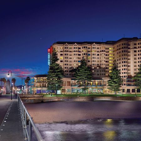 Отель Stamford Grand Adelaide Гленелг Экстерьер фото The photo shows a waterfront scene at dusk, featuring a large building lit up against a darkening sky. The architecture appears modern and prominent, with several windows illuminated. In the foreground, there is a walkway or pier with fencing along t