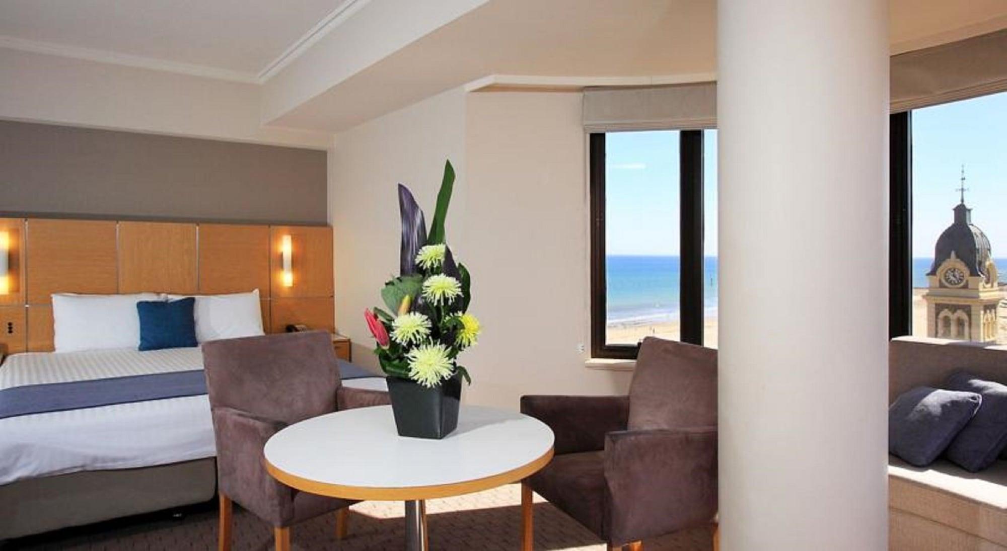 Отель Stamford Grand Adelaide Гленелг Экстерьер фото The photo shows a cozy hotel room with a comfortable seating arrangement. There are two brown chairs positioned around a round table, which features a decorative flower arrangement in a black vase. The background includes a window that offers a view 