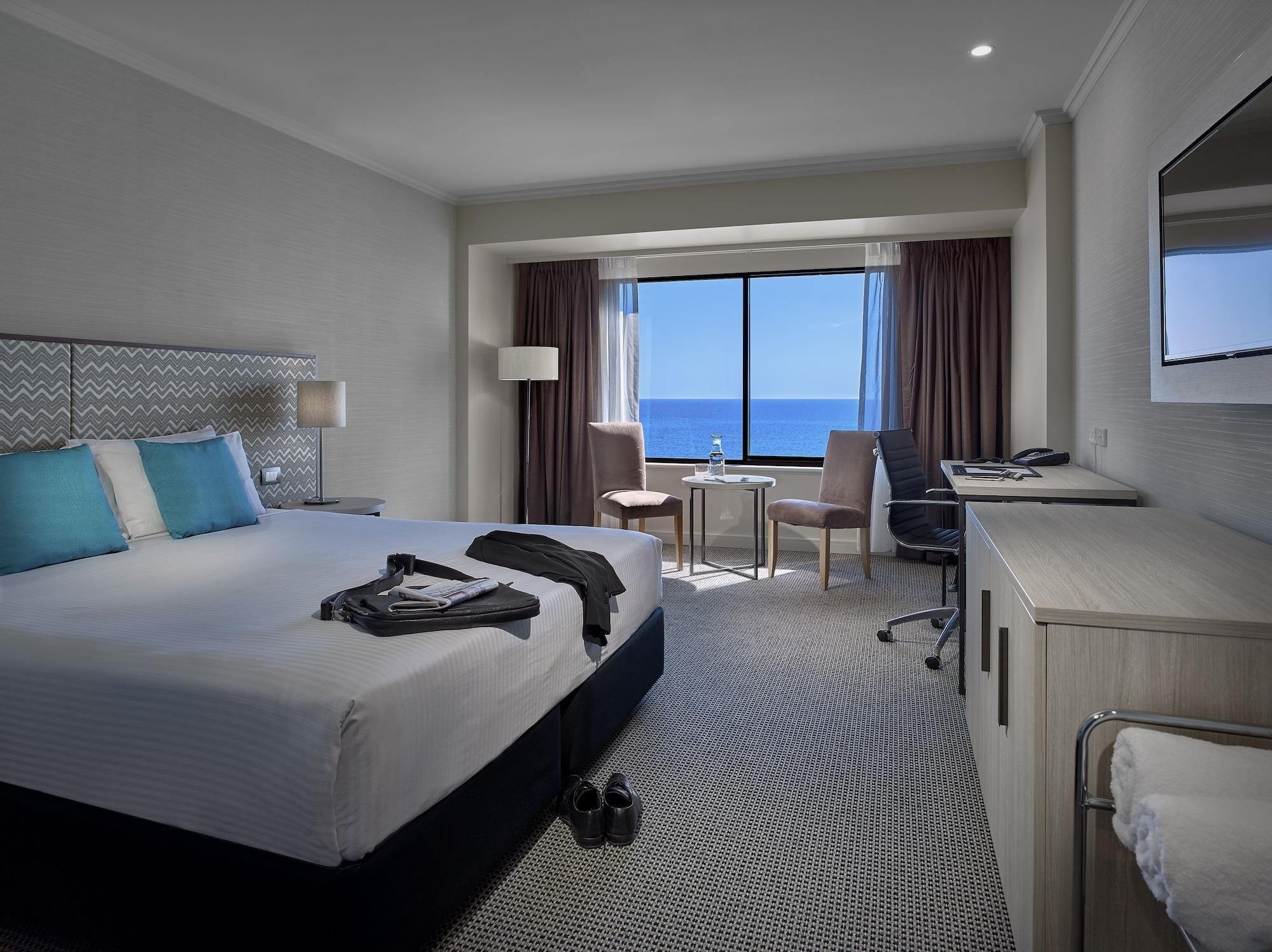 Отель Stamford Grand Adelaide Гленелг Экстерьер фото The photo shows a modern hotel room featuring a queen-sized bed with white linens and two aqua blue decorative pillows. There are bedside lamps and a large window with a view of the ocean. The room also has a small seating area with two chairs and a 