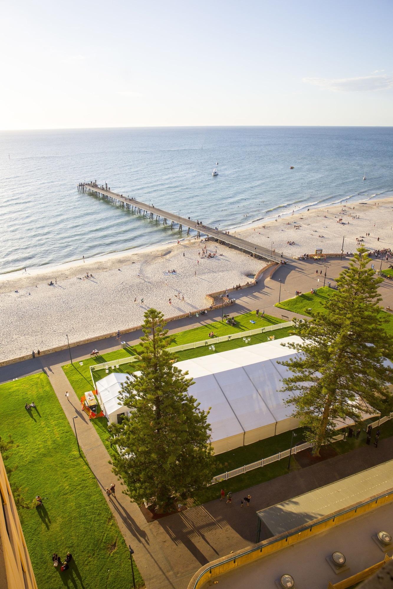 Отель Stamford Grand Adelaide Гленелг Экстерьер фото The photo shows a scenic view of a beach with a long pier extending into the water. The beach is sandy, and there are people enjoying the sun and walking along the shore. In the foreground, there is a large white tent set up on a grassy area, likely 