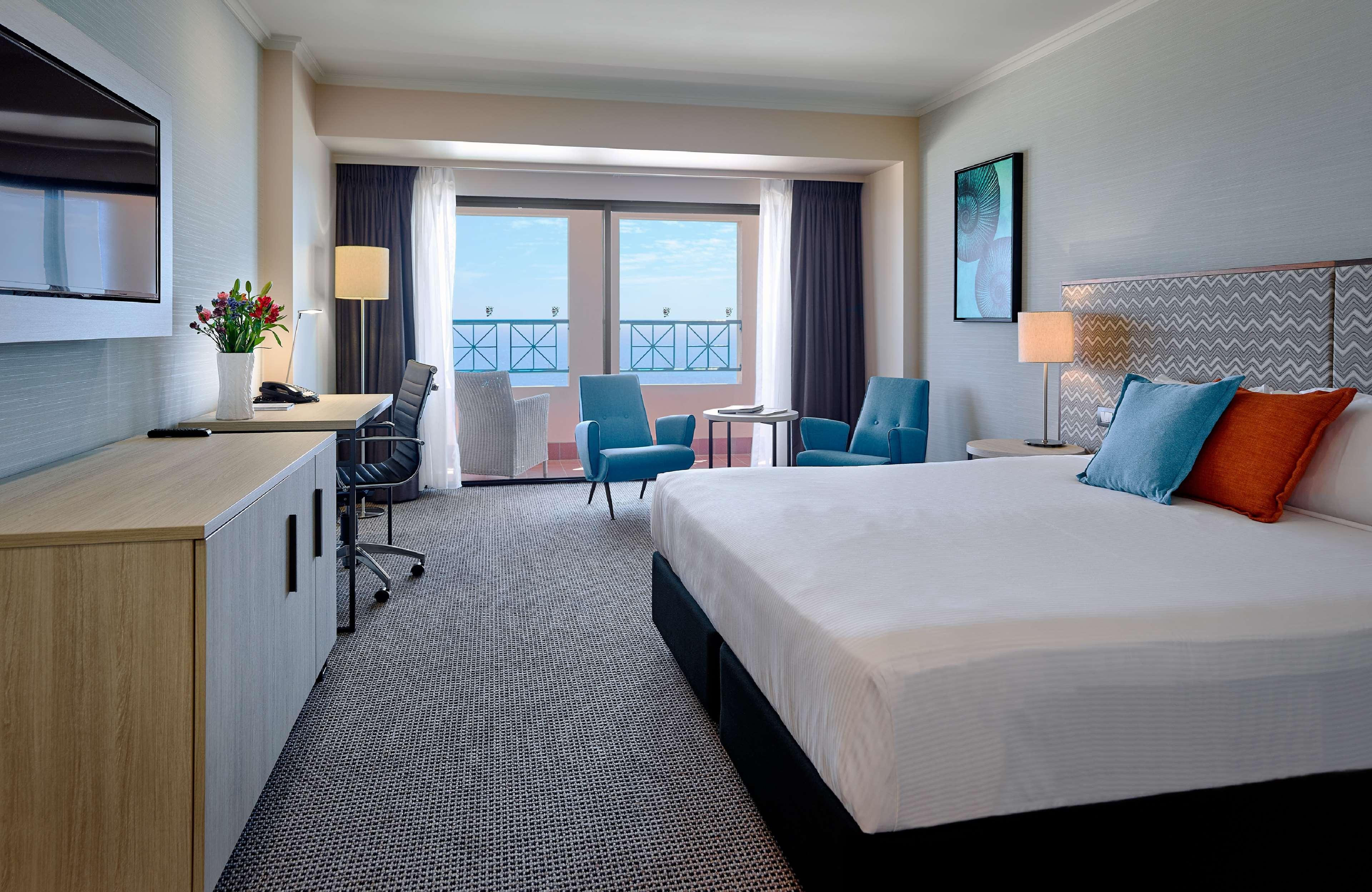 Отель Stamford Grand Adelaide Гленелг Экстерьер фото The photo depicts a modern hotel room. The room features a large bed with a white comforter, and there are decorative pillows in orange and blue. There is a flat-screen TV mounted on the wall, and a desk with an office chair is positioned near the wi