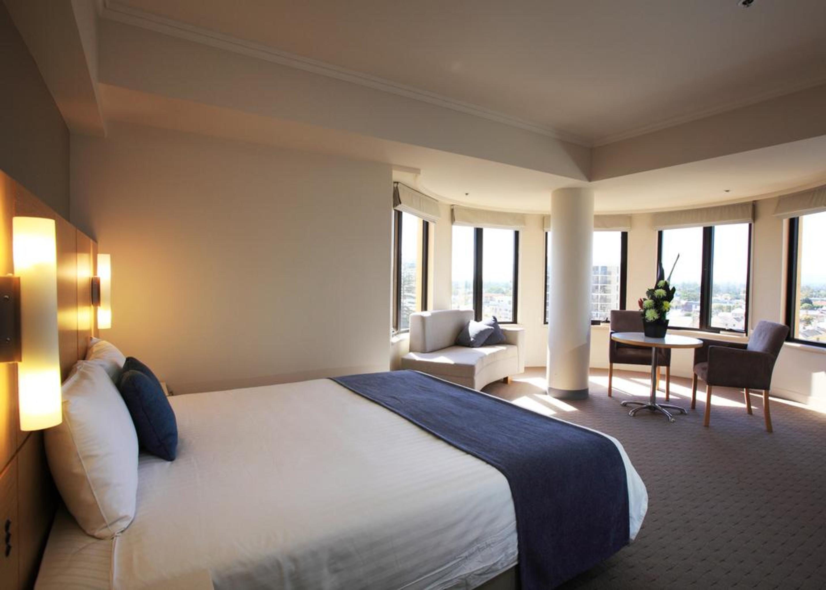 Отель Stamford Grand Adelaide Гленелг Экстерьер фото The photo shows a hotel room with a modern and spacious design. There is a large bed with white linens and a dark blue throw blanket. The room features multiple windows that allow plenty of natural light, and there is a small seating area near the wi