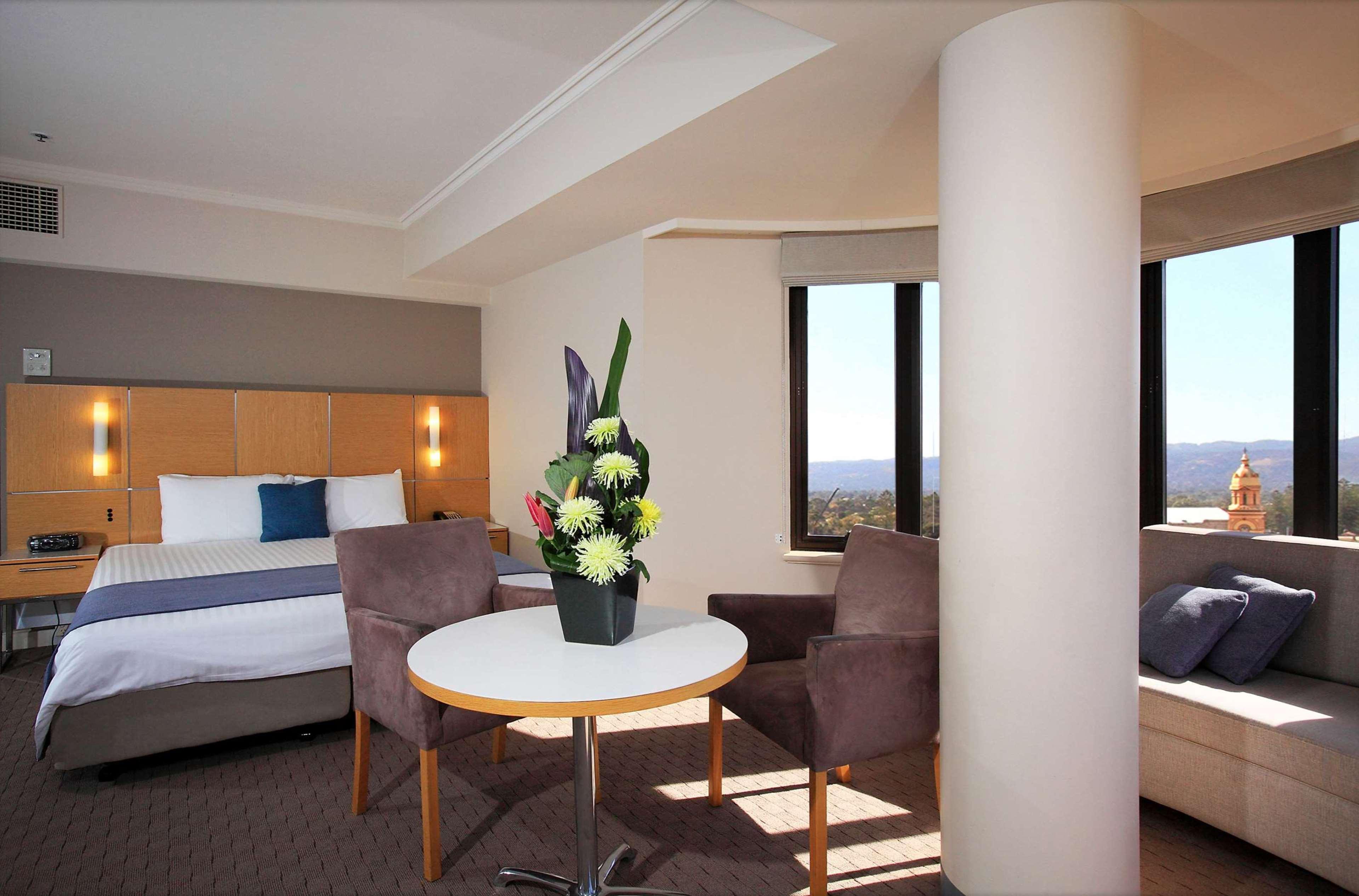 Отель Stamford Grand Adelaide Гленелг Экстерьер фото The photo shows a hotel room with a modern design. On one side, there is a plush bed with white and blue bedding. In the center of the room, there's a small round table with two gray upholstered chairs around it. A vase of flowers sits on the table, 