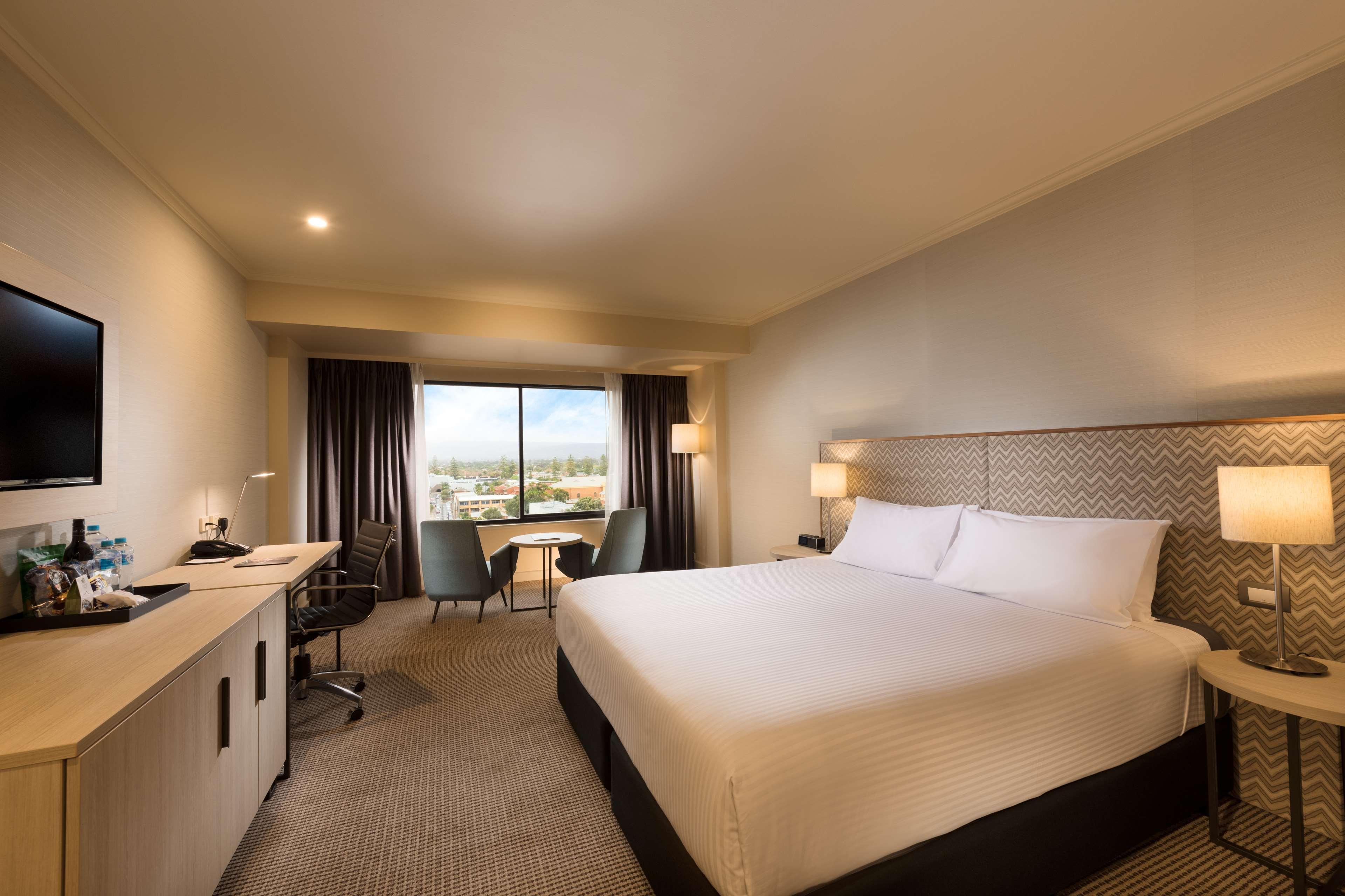 Отель Stamford Grand Adelaide Гленелг Экстерьер фото The photo shows a modern hotel room. It features a large bed with white linens, a desk with a chair, and a flat-screen TV mounted on the wall. There are two armchairs and a small table near a large window that lets in natural light, providing a view 