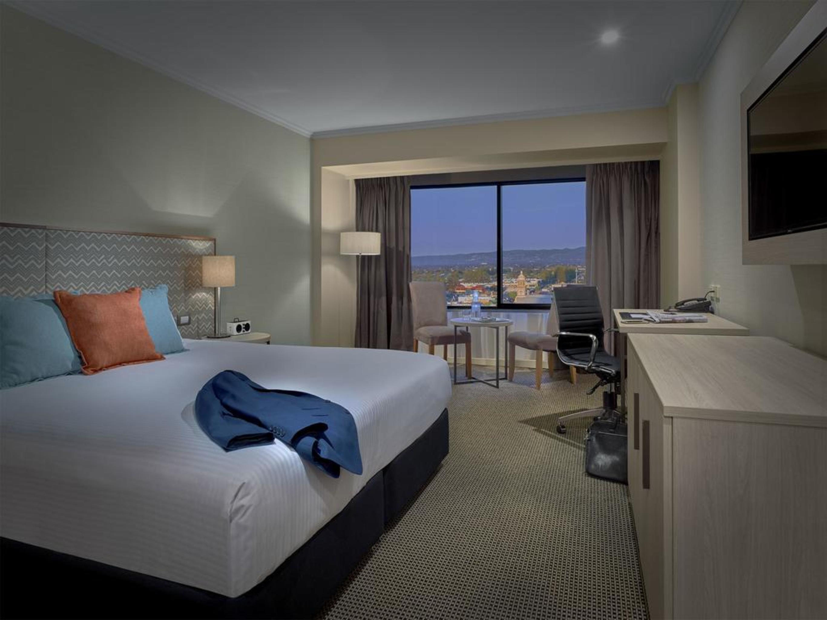Отель Stamford Grand Adelaide Гленелг Экстерьер фото The photo shows a modern hotel room. It features a large double bed with white bedding and decorative pillows in orange and blue. There's a nightstand with lamps on either side of the bed. A large window overlooks a view outside, and there's a desk a