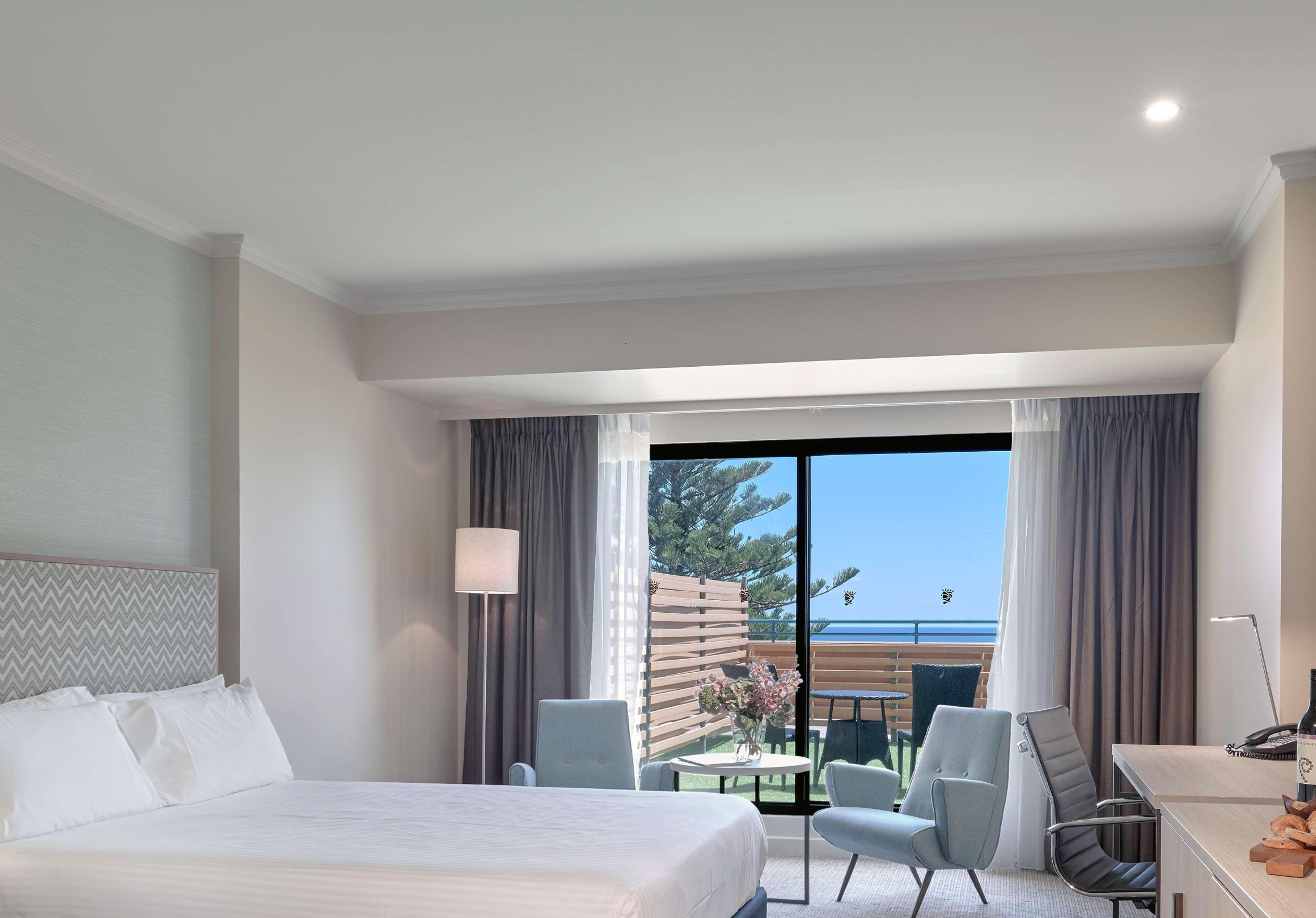 Отель Stamford Grand Adelaide Гленелг Экстерьер фото The photo shows a modern hotel room featuring a large bed with white bedding. There are two armchairs positioned near a window that offers a view of an outdoor area, possibly a balcony or patio with greenery. The room has light-colored walls and curt