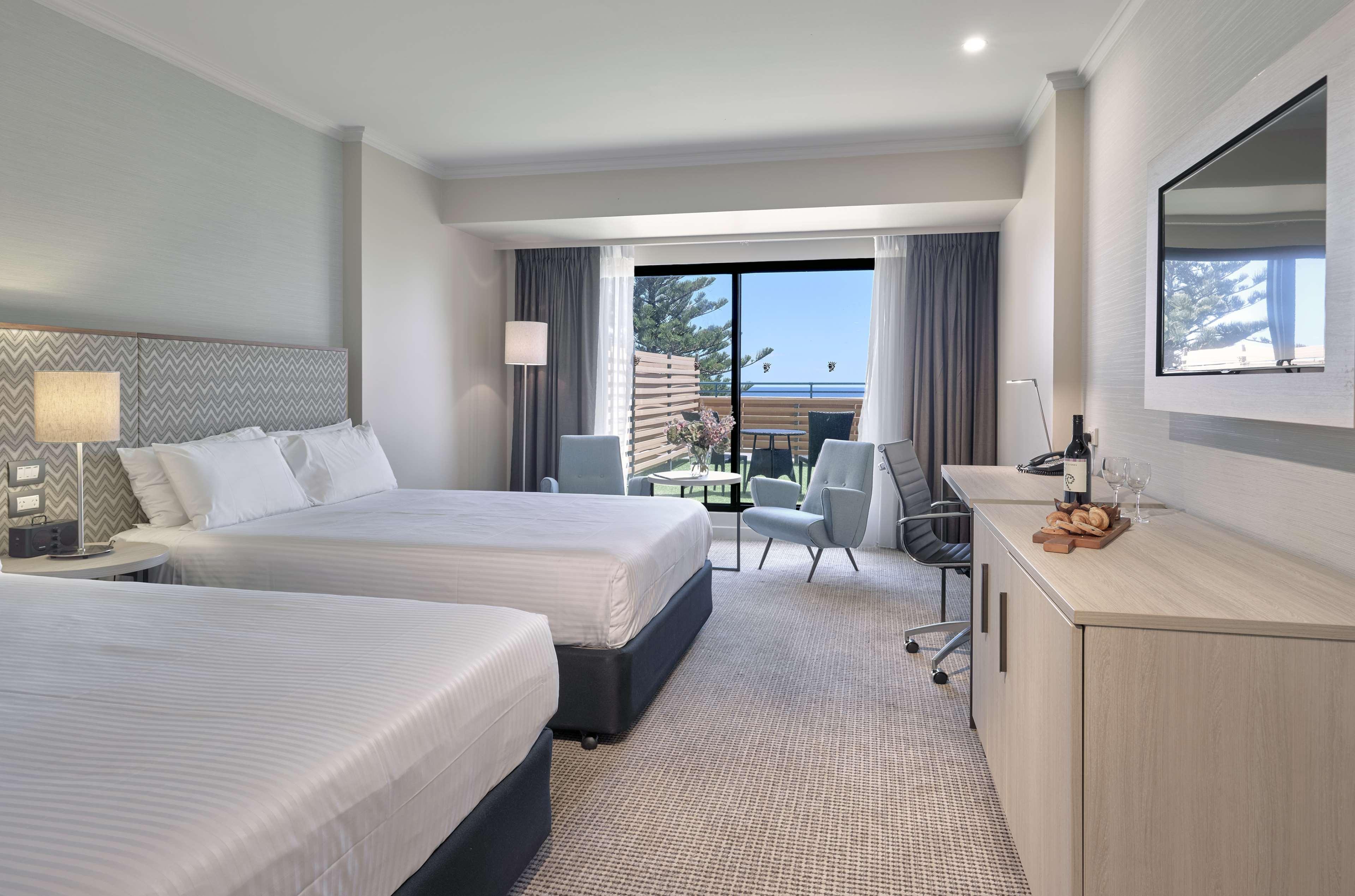 Отель Stamford Grand Adelaide Гленелг Экстерьер фото The photo shows a modern hotel room featuring two beds with white linens. There's a large window that provides natural light and a view of a balcony area. The room includes two chairs and a small table, creating a cozy sitting area. A television is m