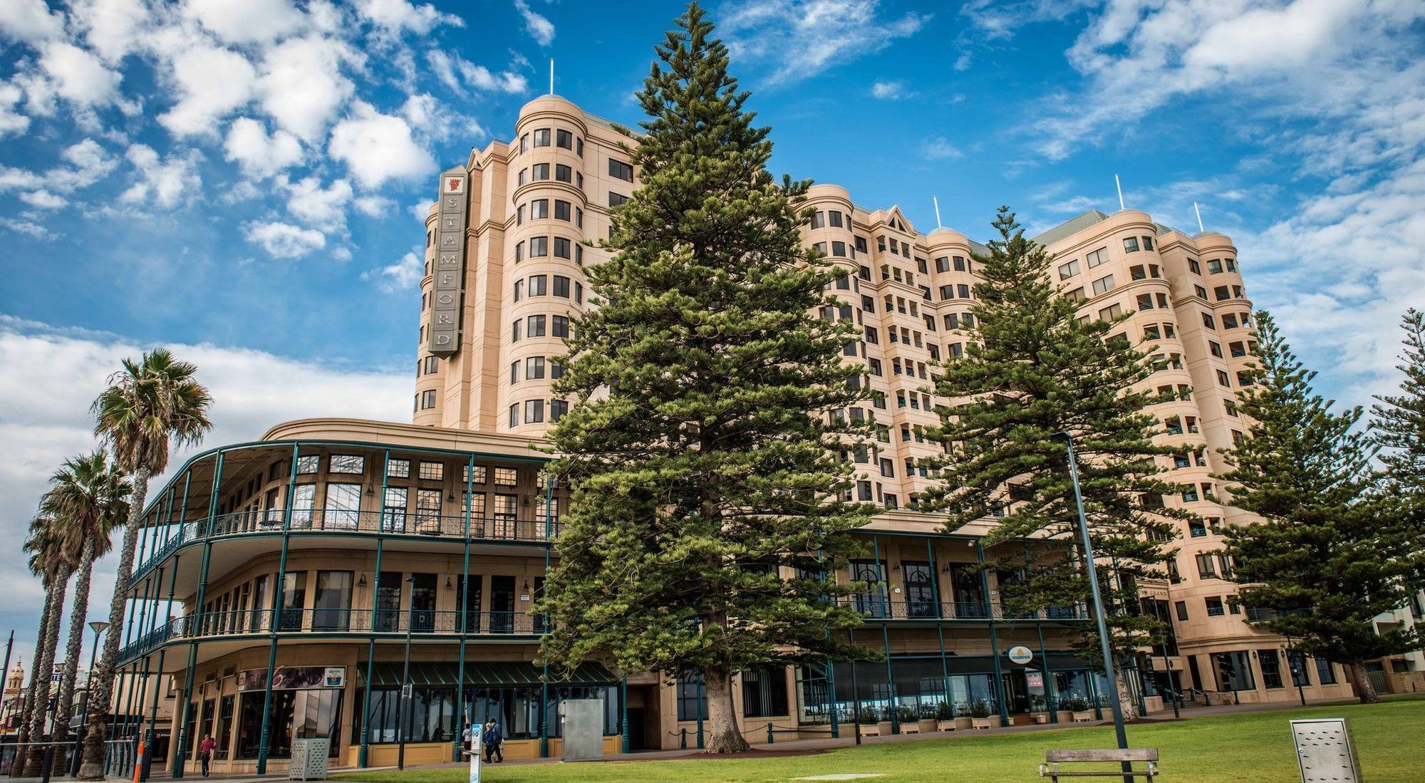 Отель Stamford Grand Adelaide Гленелг Экстерьер фото The image shows a large building with a distinctive architectural style, characterized by a tan or beige color. The structure appears to have multiple floors and rounded edges. In front of the building, there are several tall green trees, which add t