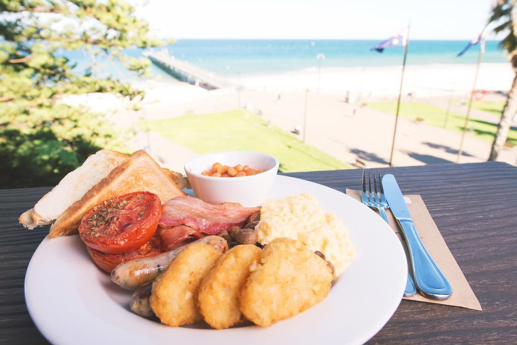 Отель Stamford Grand Adelaide Гленелг Экстерьер фото The photo shows a plate of breakfast food set against a beachside backdrop. The plate includes several items: crispy bacon, a grilled tomato, sausage links, golden hash browns, and a small serving of baked beans. There is also a piece of toast on the
